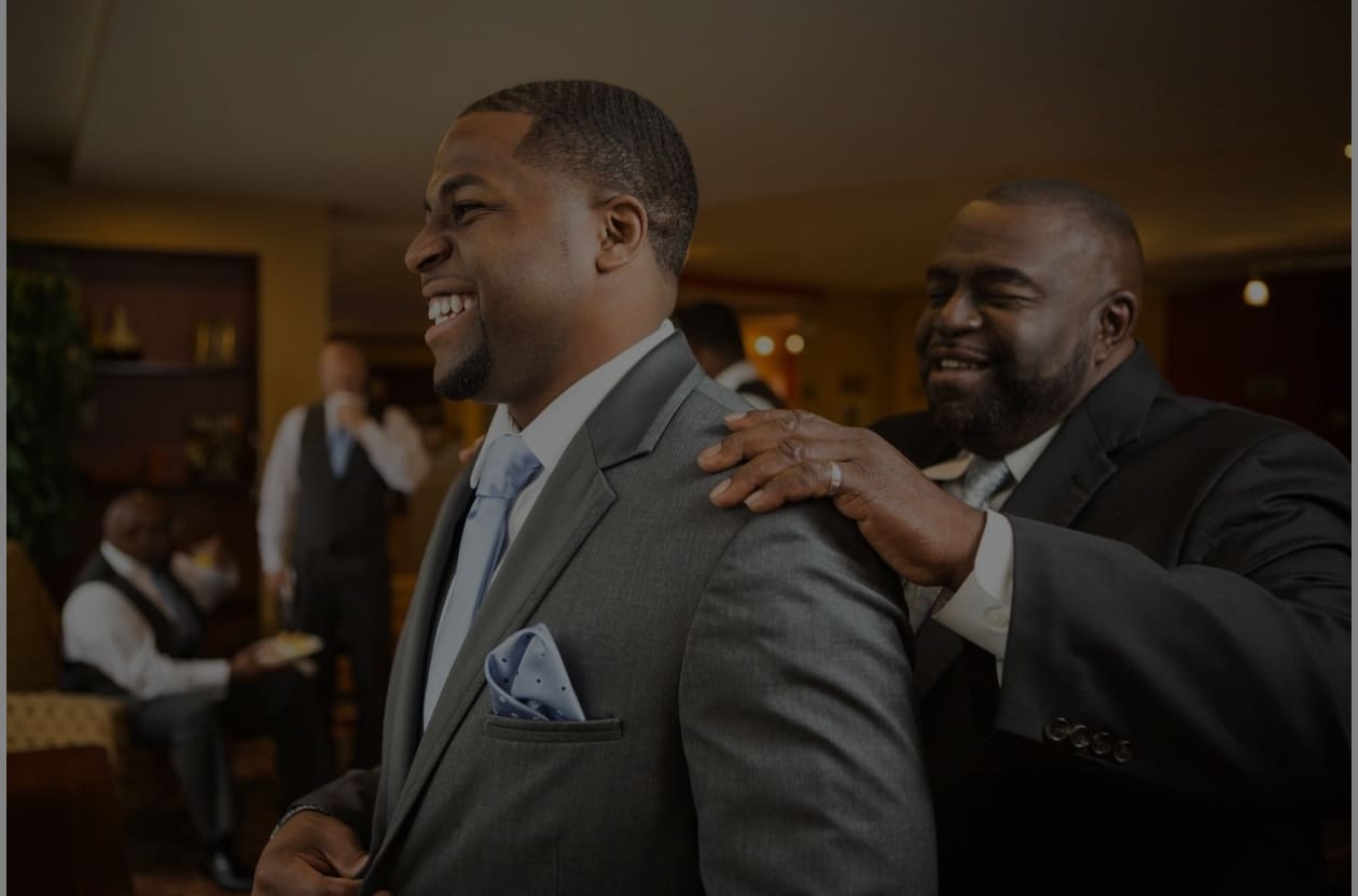 A man in suit and tie getting help from another man.